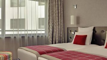 Una habitación de hotel con dos camas, una mesita de noche, una lámpara y un ventanal con cortinas.