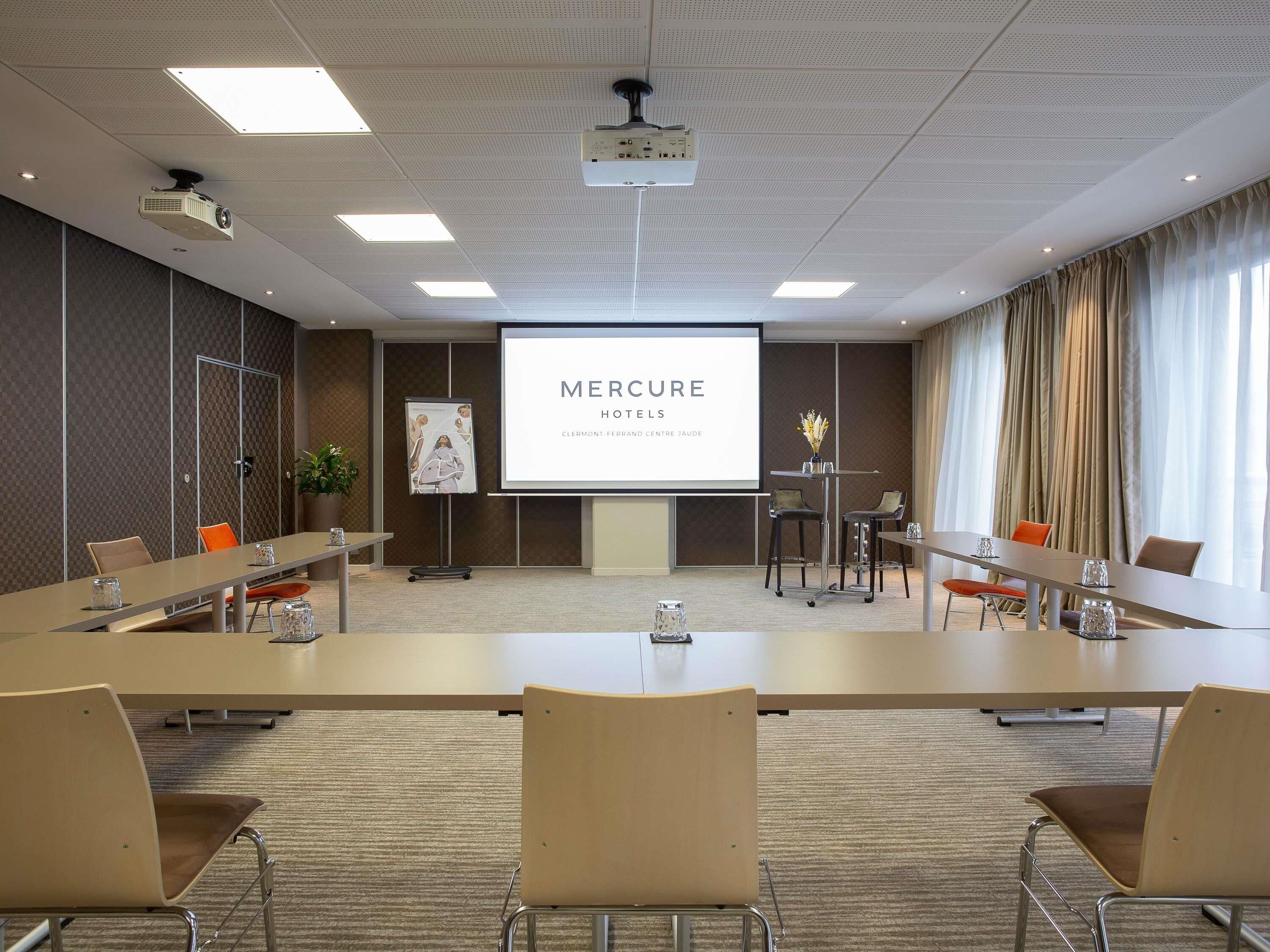 A conference room with a large table, chairs, a projector, and a presentation screen displaying 'MERCURE HOTELS'.