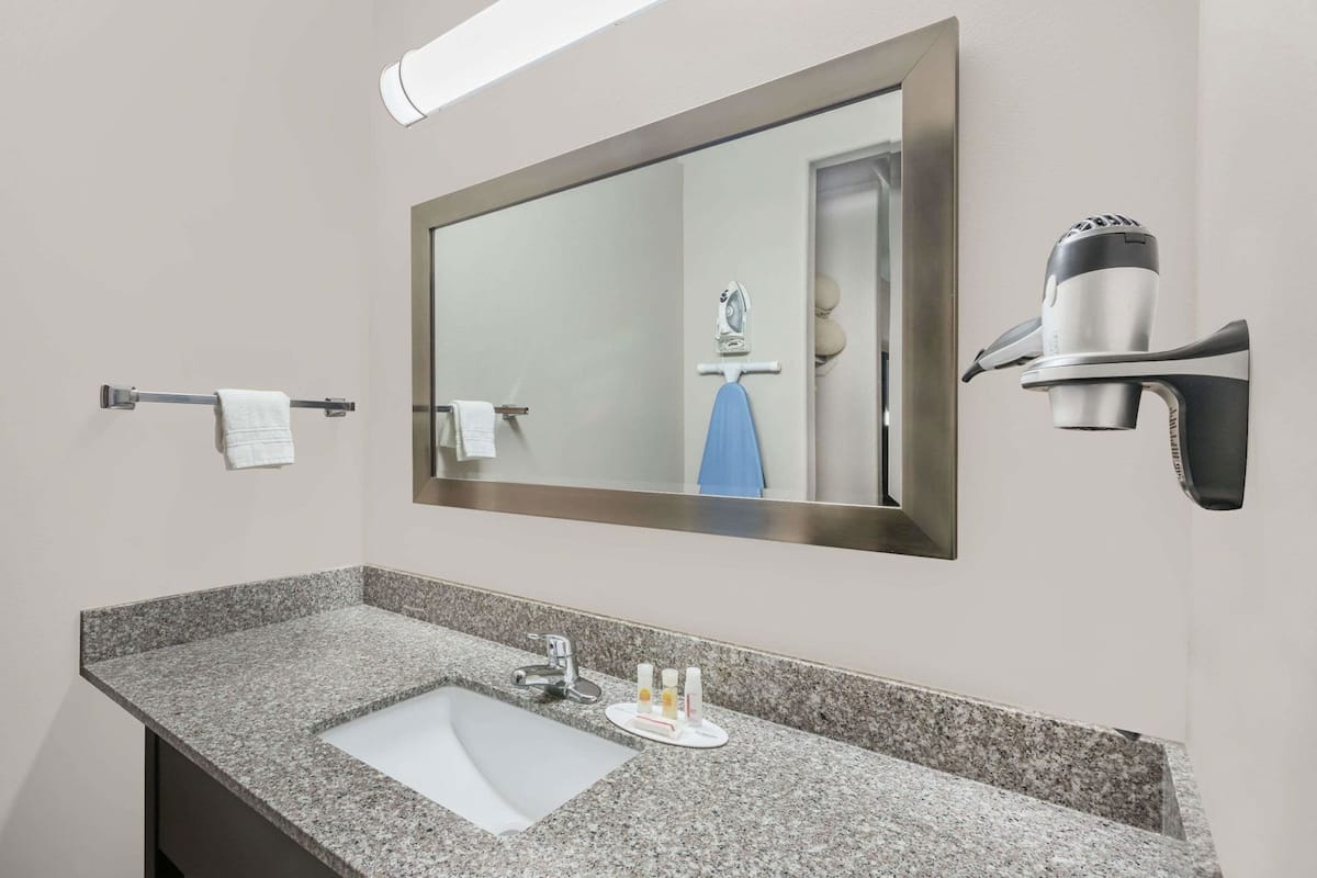 A bathroom with a granite countertop sink, a mirror with a towel rack, and a hairdryer mounted on the wall.