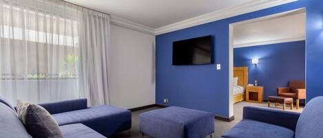 Una habitación de hotel con una cama grande, un televisor montado en la pared y un área de descanso con sofás y otomanas.