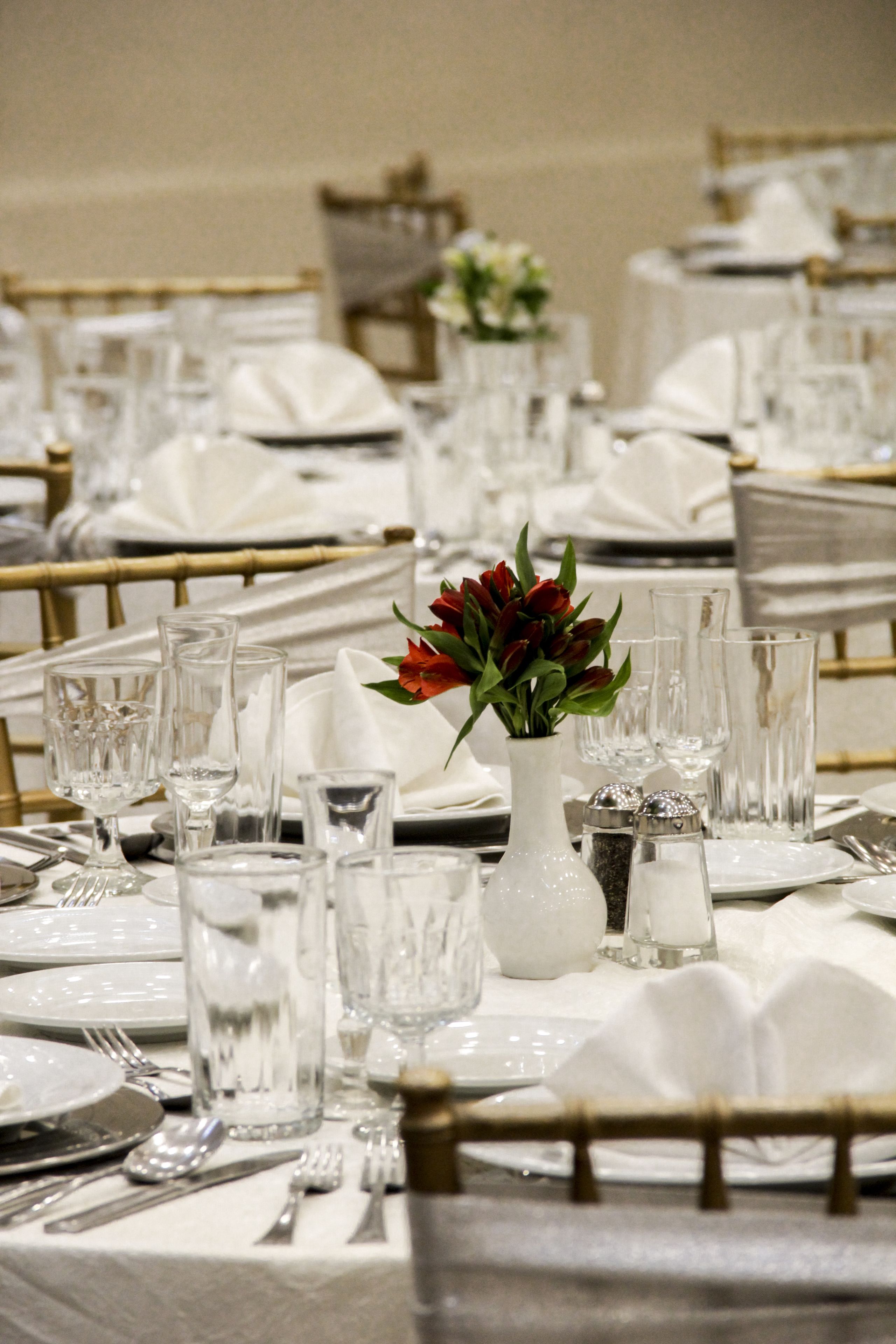 A banquet table set with white tablecloths, glassware, and a centerpiece with red flowers.