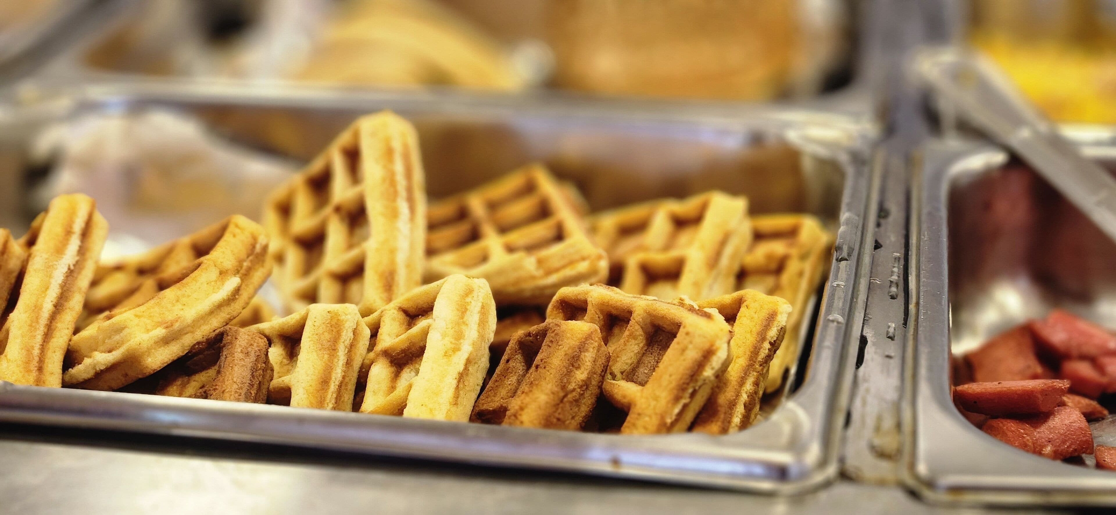 A close-up of a waffle iron with waffles and a container of sausages.