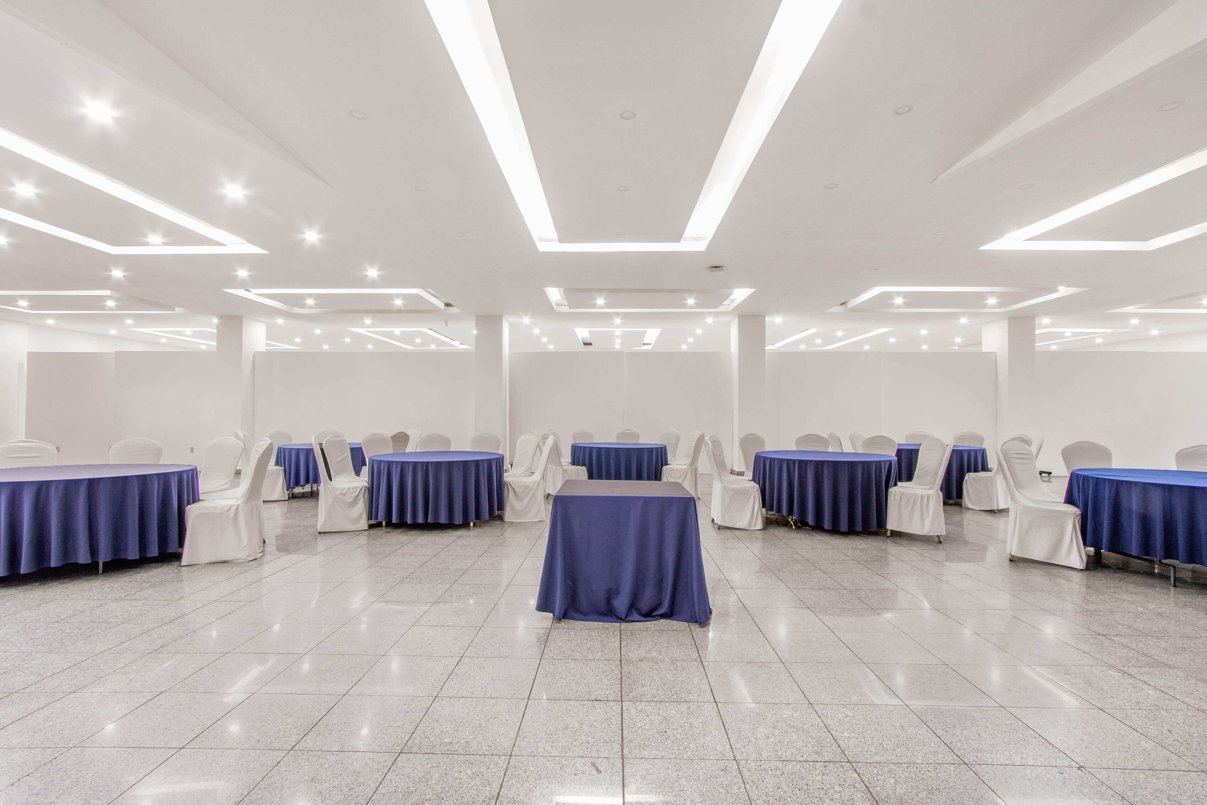Un salón de conferencias con varias mesas y sillas, paredes blancas y piso de baldosas.