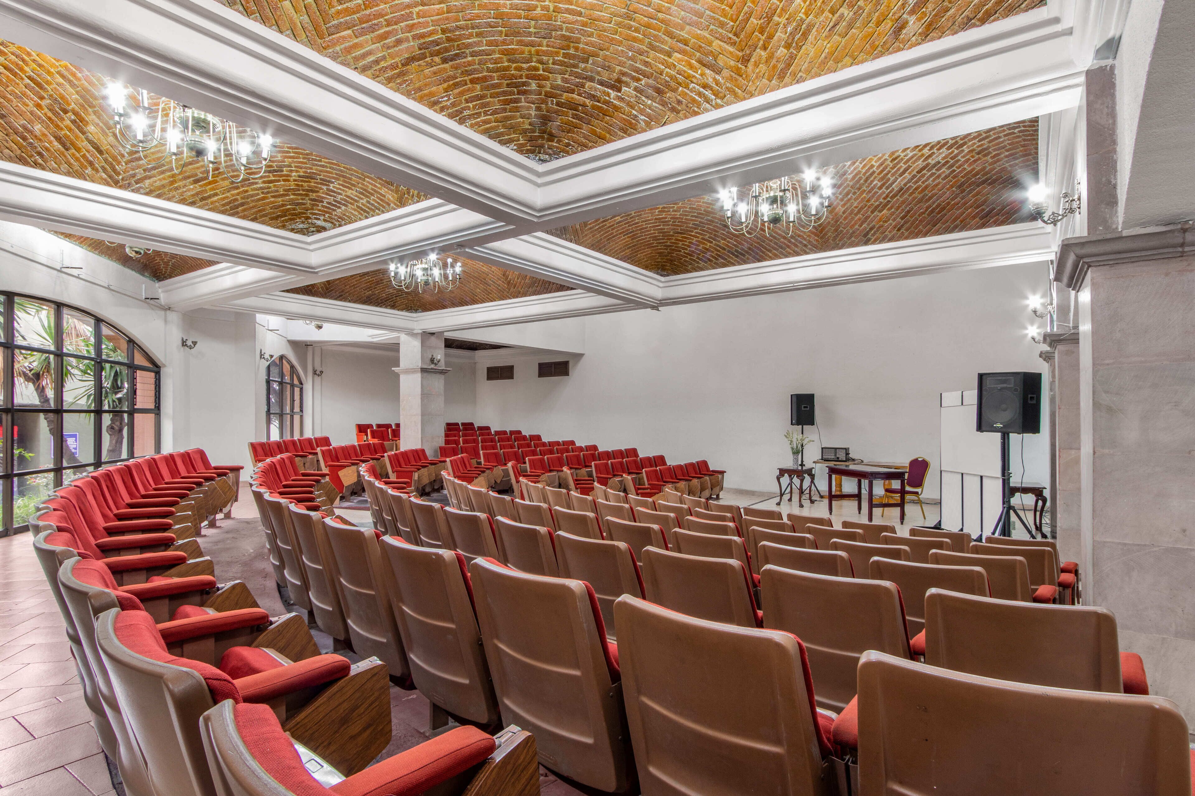 A conference room with rows of chairs, a stage with a speaker, and a brick ceiling.