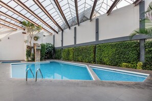 An indoor swimming pool with a glass ceiling, surrounded by greenery and a tiled floor.