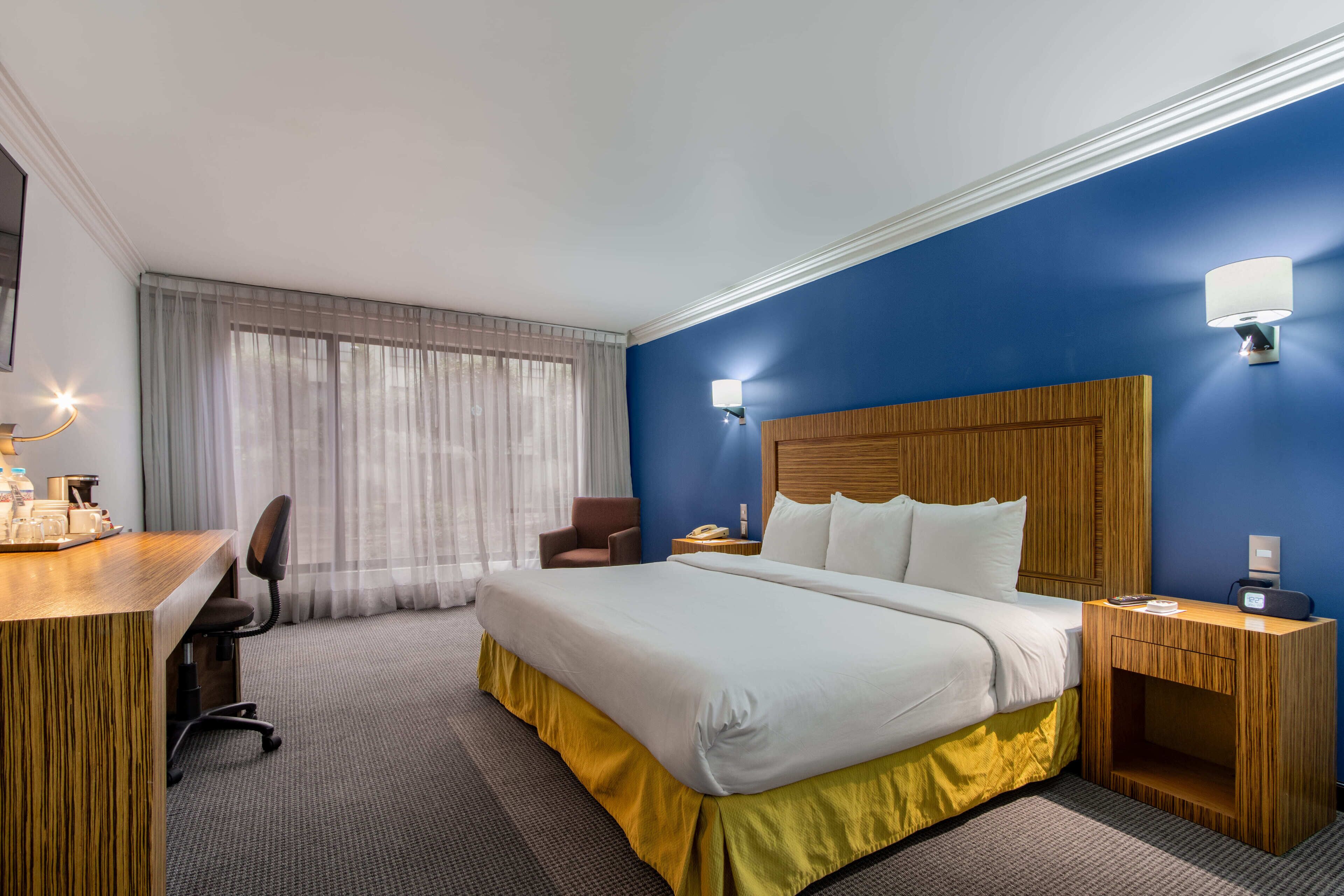 Habitación de hotel con una cama grande, un escritorio con silla, una mesita de noche de madera y una lámpara de pared.