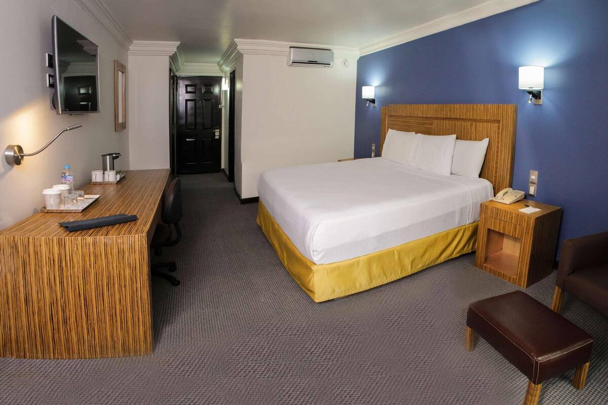 A hotel room with a large bed, bedside table, desk, chair, and a small wooden stool.