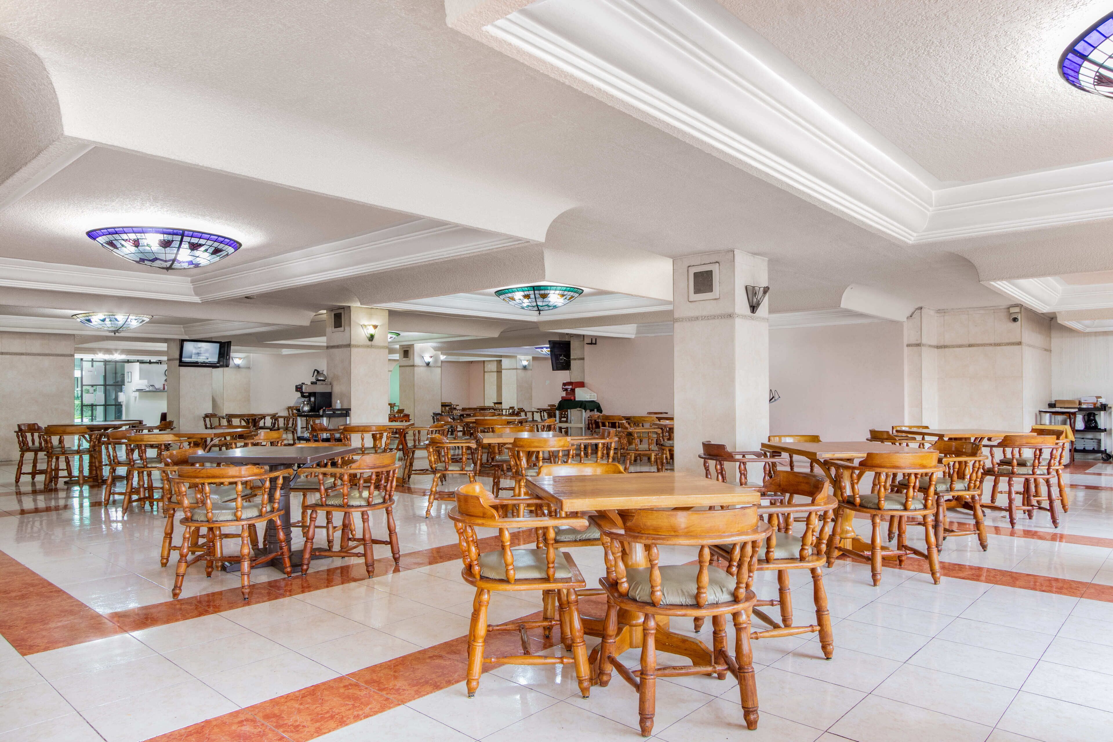 A spacious dining area with numerous wooden tables and chairs, a high ceiling with decorative lighting, and a clean, tiled floor.