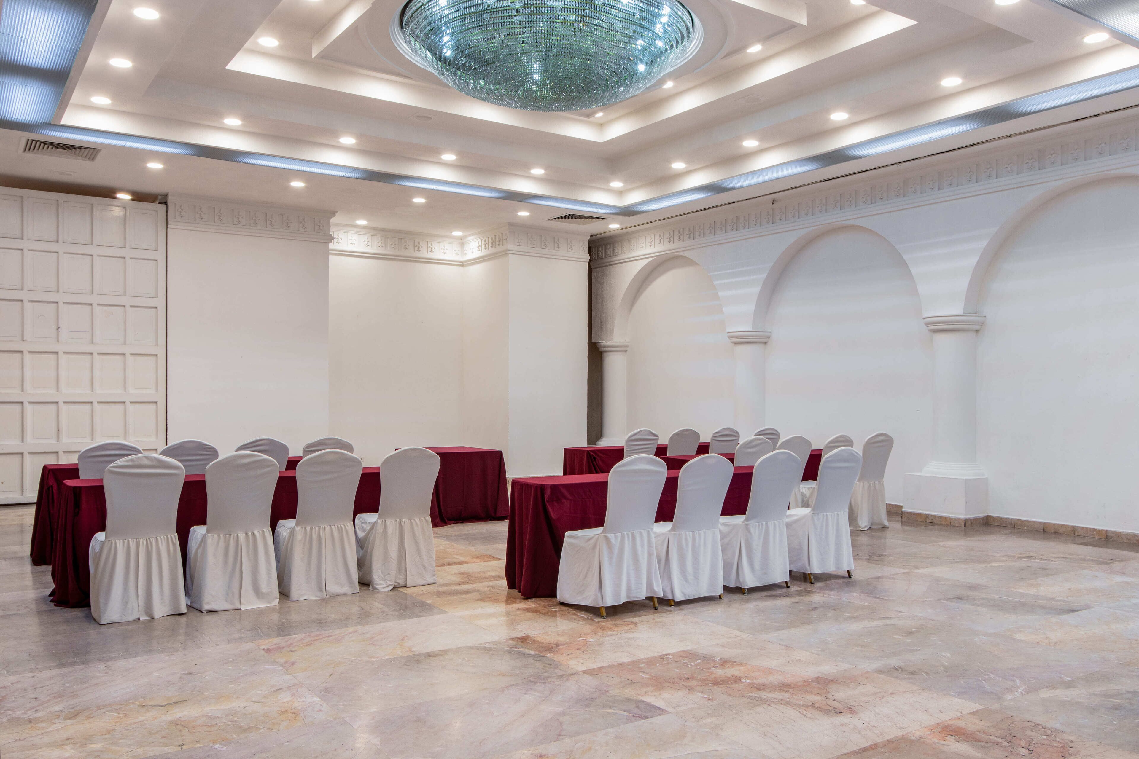 Un salón de conferencias con suelo de mármol, sillas blancas con fundas blancas, manteles rojos y una lámpara de araña grande.