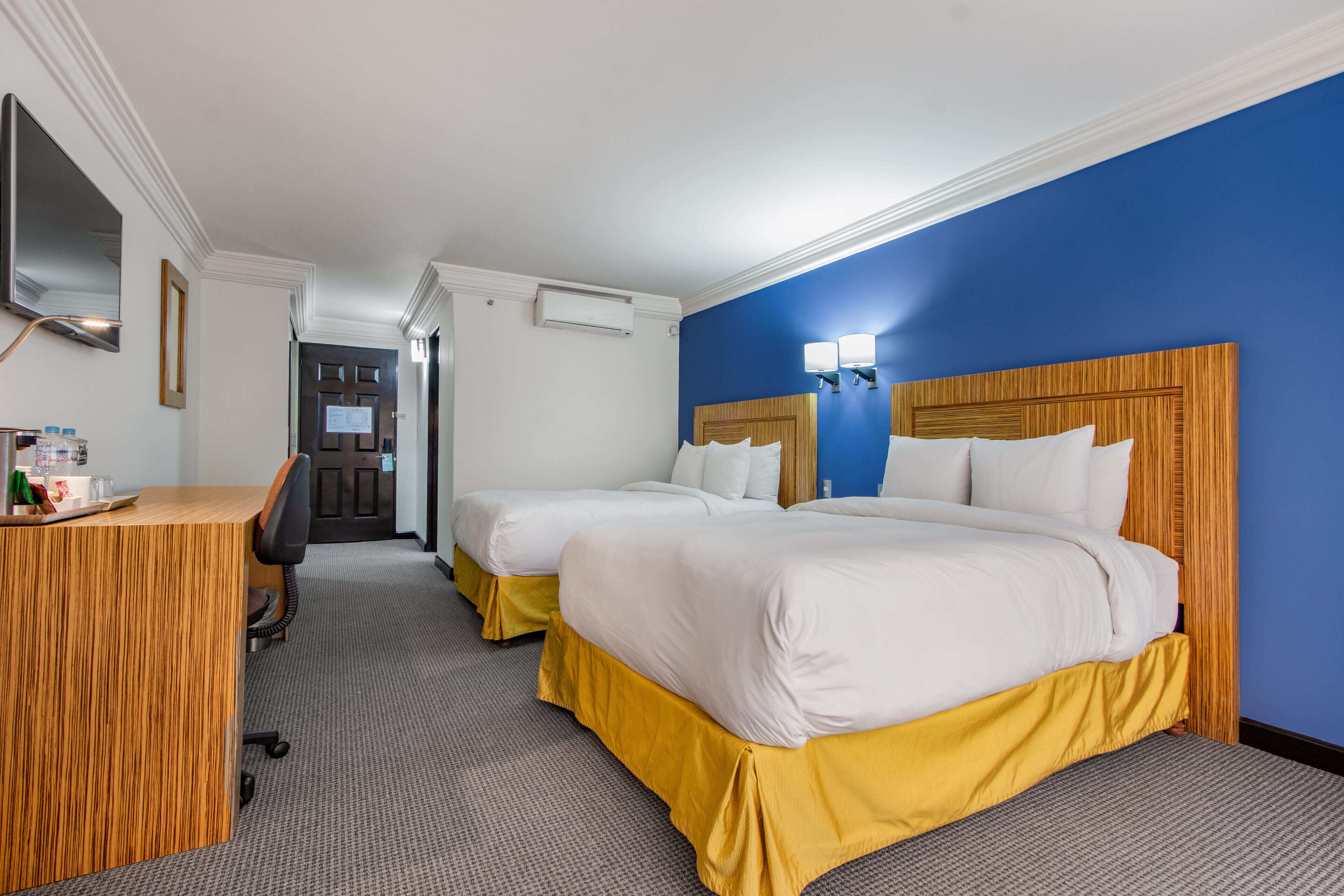 Habitación de hotel con dos camas, un escritorio, un espejo y una puerta que da a un pasillo.