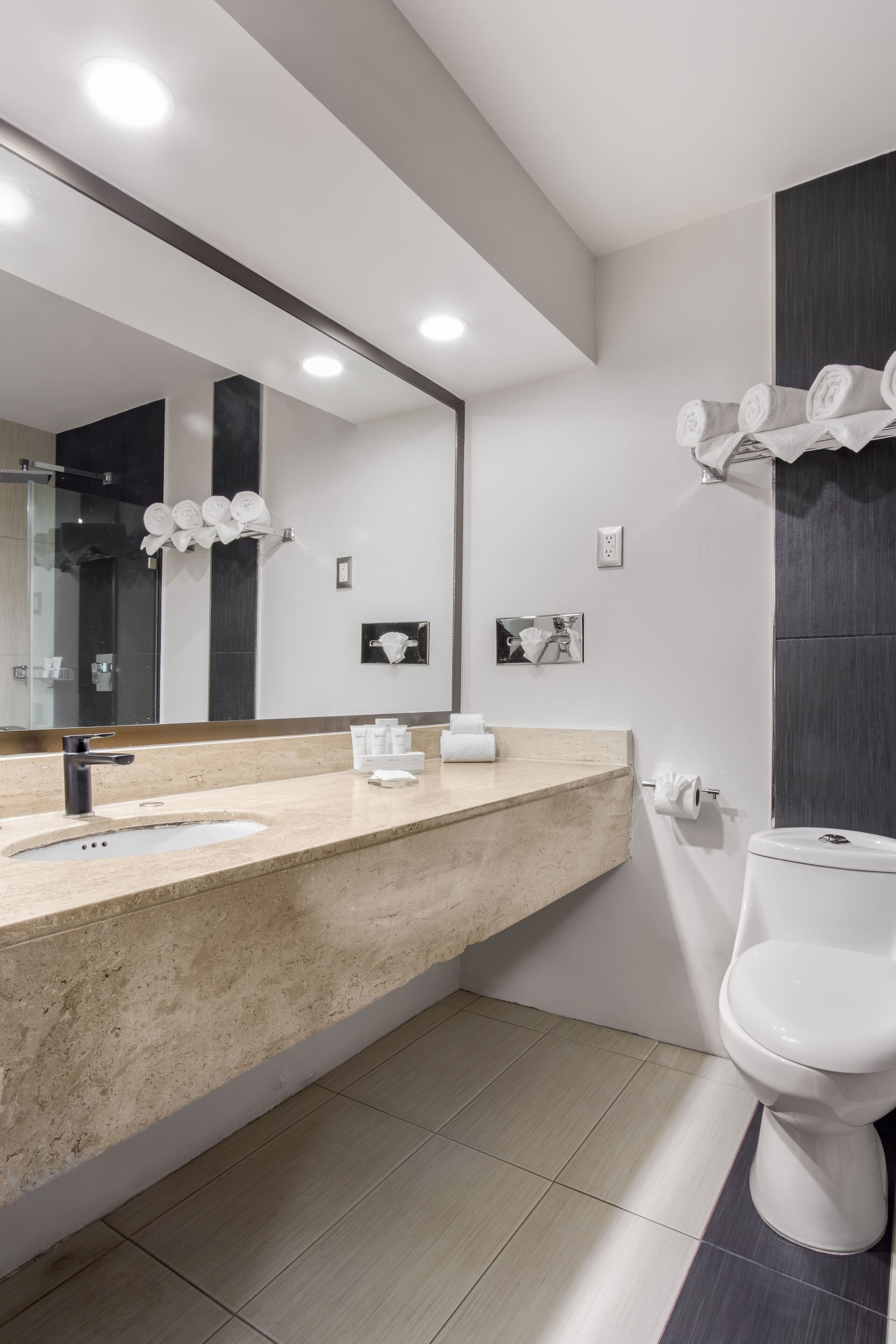 A modern bathroom with a beige countertop, a large mirror, a white toilet, and a towel rack with neatly folded towels.