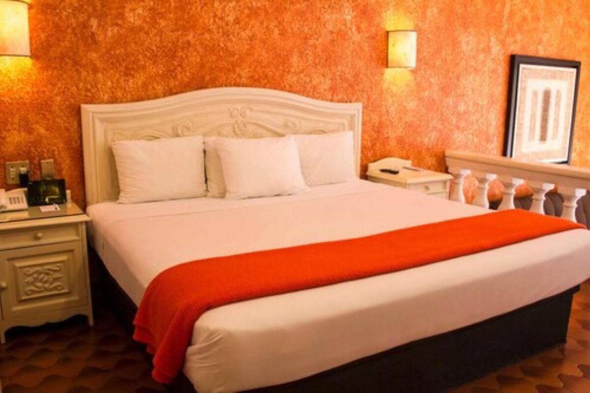A hotel room with a white bed, a red throw blanket, a nightstand with a phone, and a wall with a textured orange pattern.