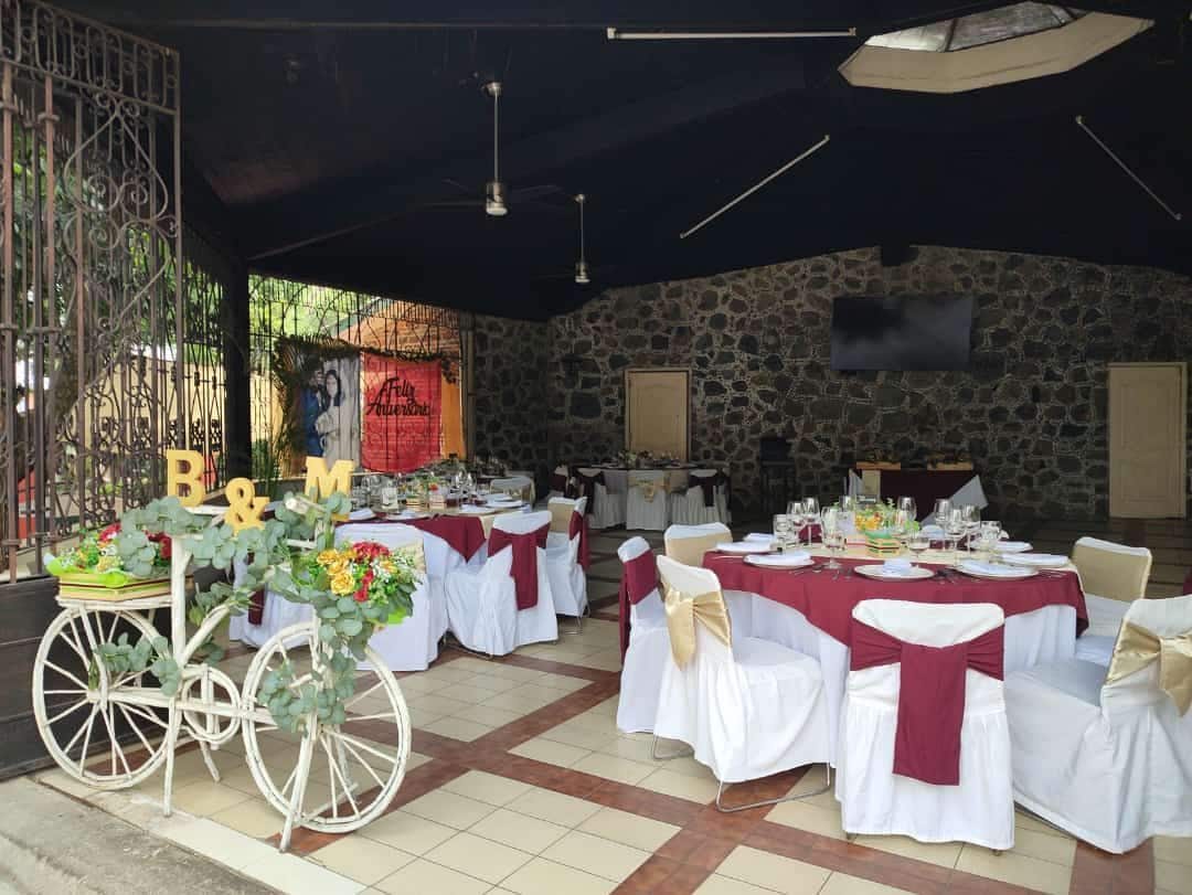 Un área de comedor al aire libre con mesas redondas cubiertas con manteles blancos y fajines rojos, frente a un fondo de pared de piedra.