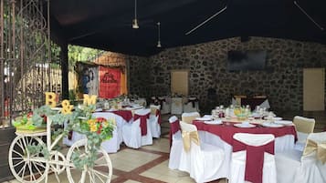 Un área de comedor al aire libre con mesas redondas cubiertas con manteles blancos y fajines rojos, frente a un fondo de pared de piedra.