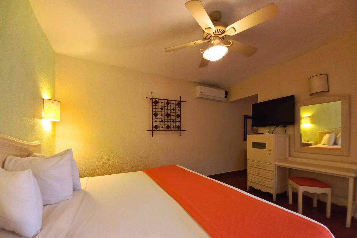 A hotel room with a bed, a desk, a chair, a ceiling fan, and a TV.