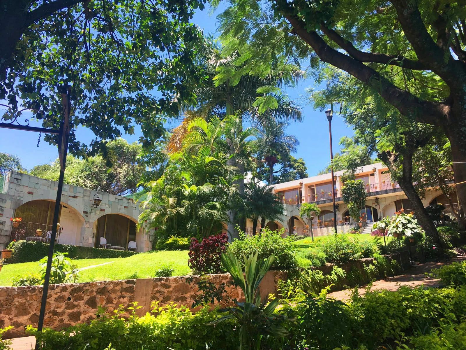 Un patio con muro de piedra, abundante follaje y un edificio con puertas arqueadas.