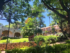 A courtyard with a stone wall, lush greenery, and a building with arched doorways. - Hotel Villa del Conquistador (Cuernavaca)