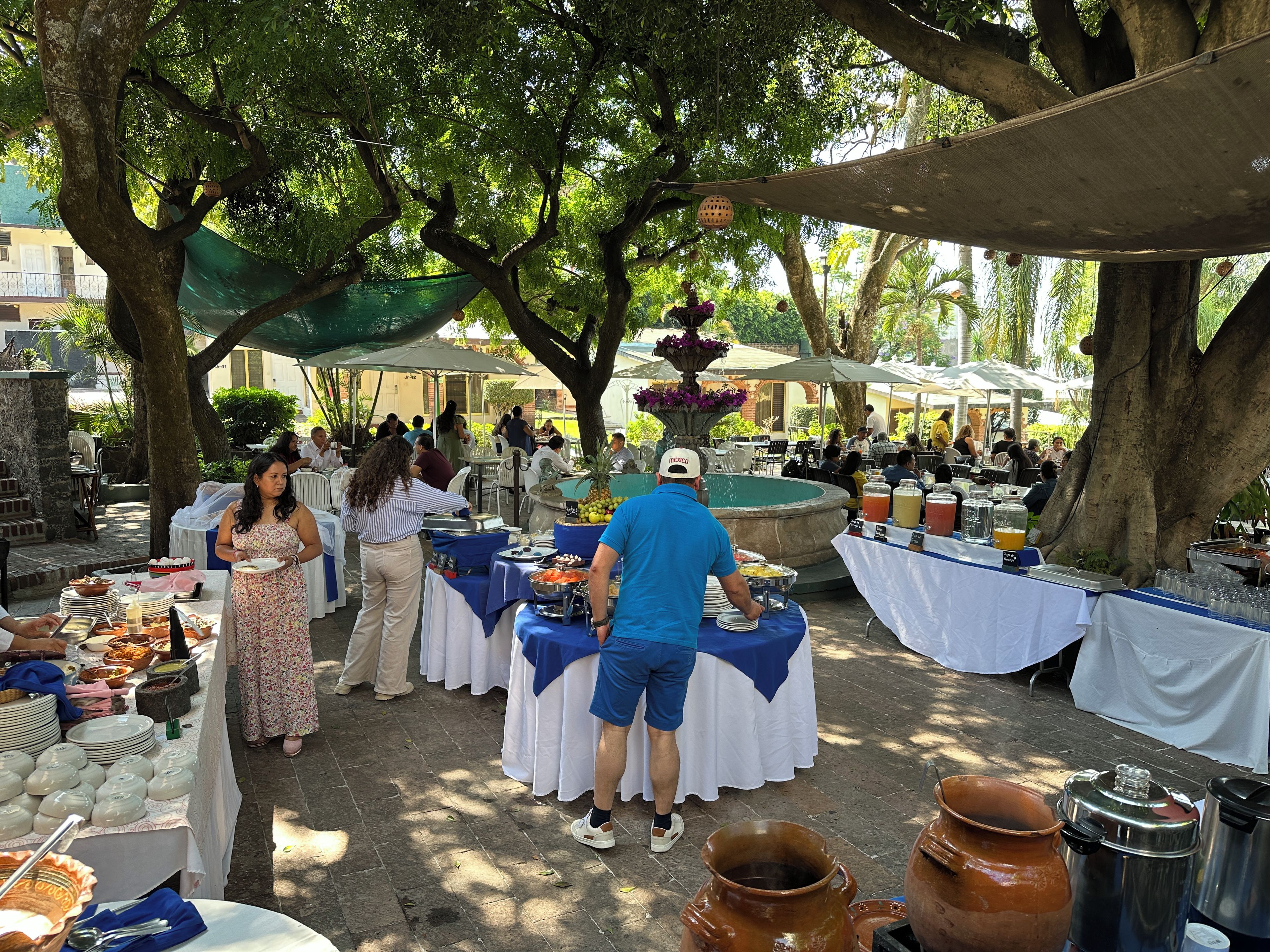 Evento al aire libre con mesas dispuestas bajo árboles, ofreciendo comida y bebidas.