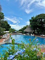 Una piscina con sillas de descanso y sombrillas, rodeada de árboles y una estructura con techo de paja.