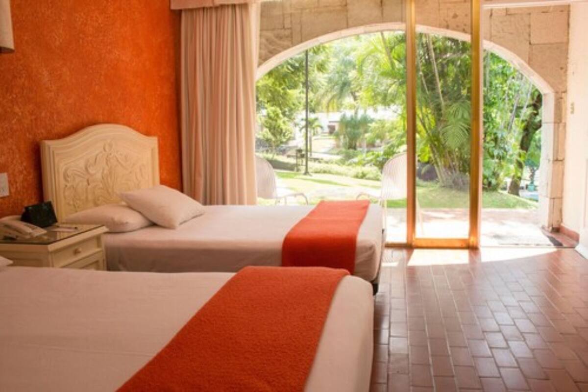 A hotel room with two beds, a nightstand with a phone, and a view of a garden through an arched doorway.