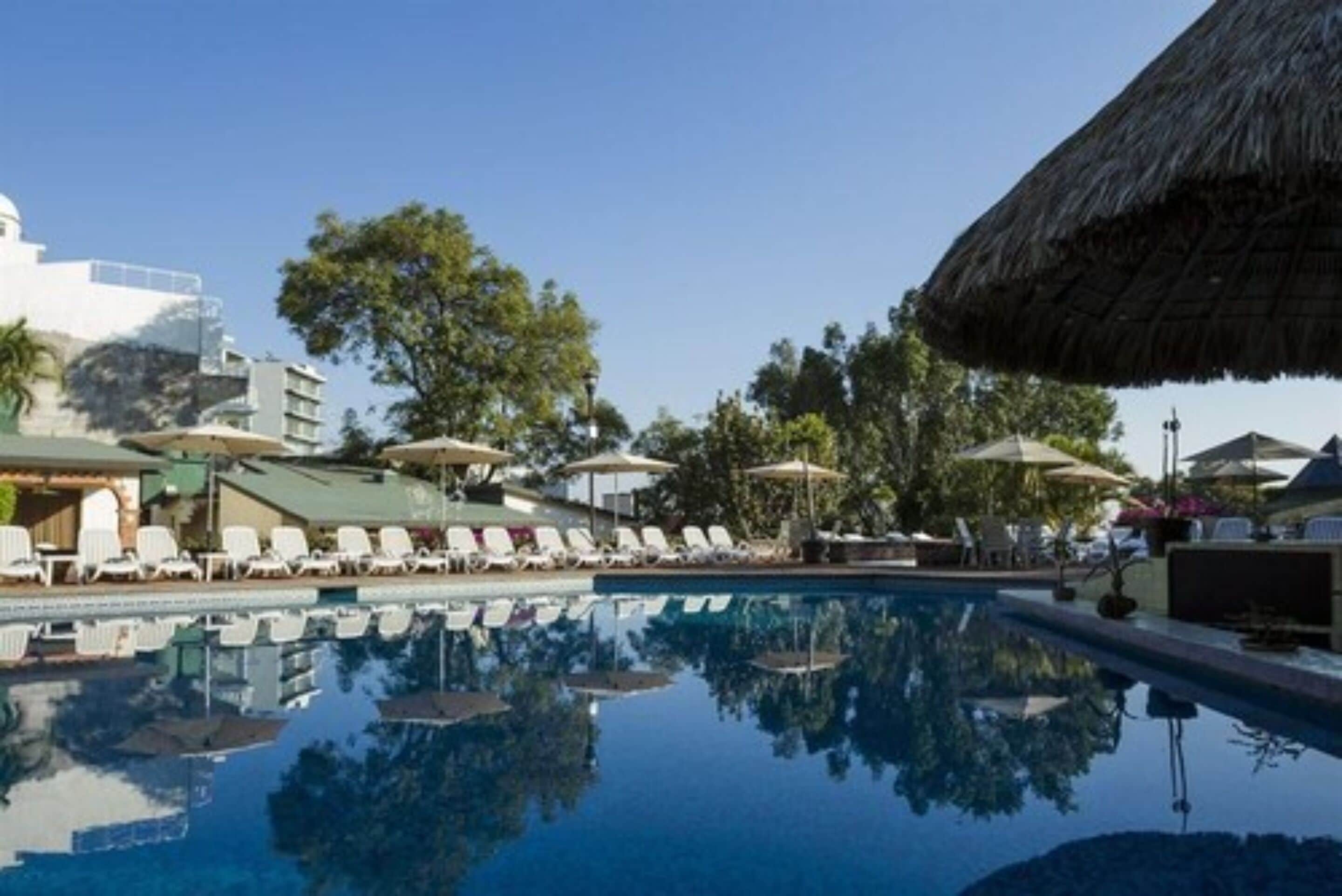 Una piscina con tumbonas y sombrillas, rodeada de edificios y árboles.