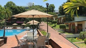 Zona junto a la piscina con sillas de descanso y sombrillas, un camino de ladrillo y abundante vegetación.