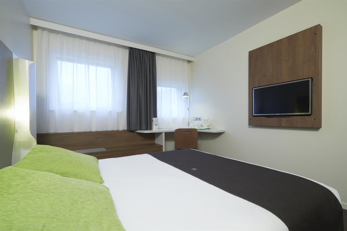 Une chambre d’hôtel avec un lit, un bureau équipé d’un ordinateur, un téléviseur fixé au mur et une fenêtre avec des rideaux.
