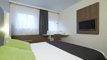 A hotel room with a bed, a desk with a computer, a TV mounted on the wall, and a window with curtains.