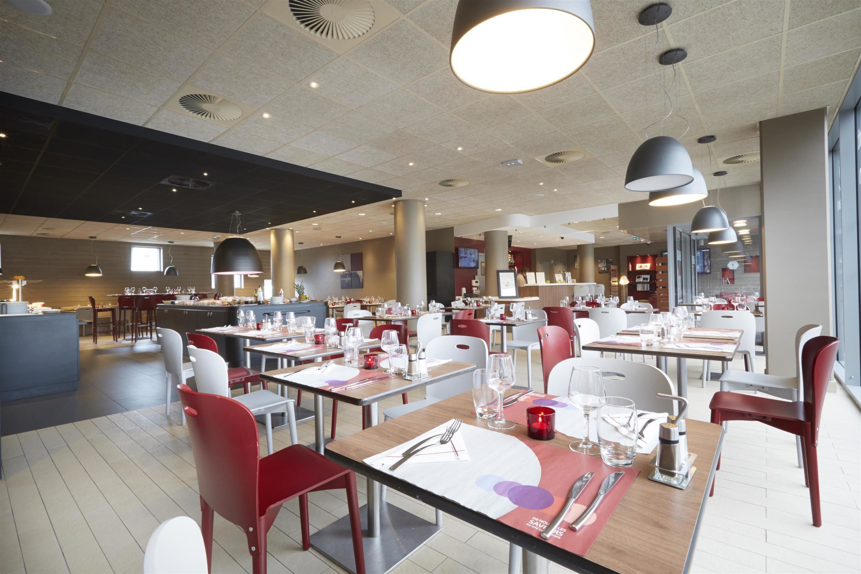 A modern restaurant with multiple dining tables, red and white chairs, and pendant lighting.