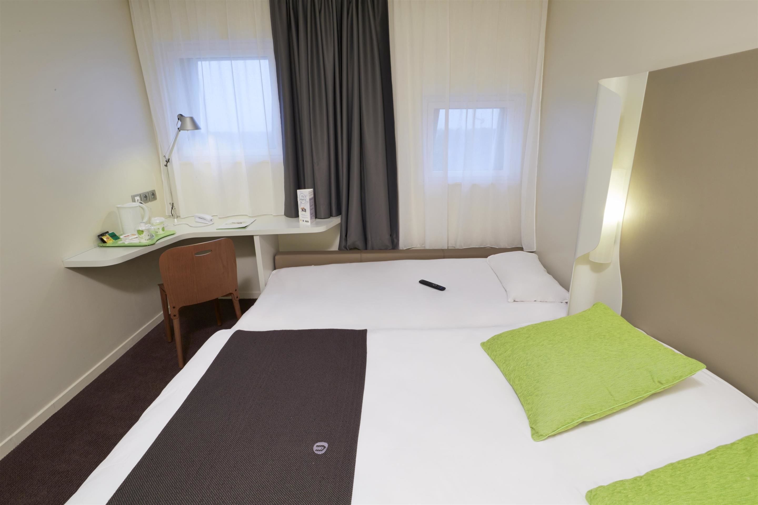 Habitación de hotel con cama, escritorio, silla, lámpara y ventana con cortinas.