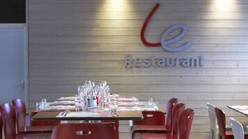 Interior de restaurante con mesas dispuestas para comer, una pared de madera con el nombre 'Le Restaurant' iluminado y sillas rojas alrededor de las mesas.