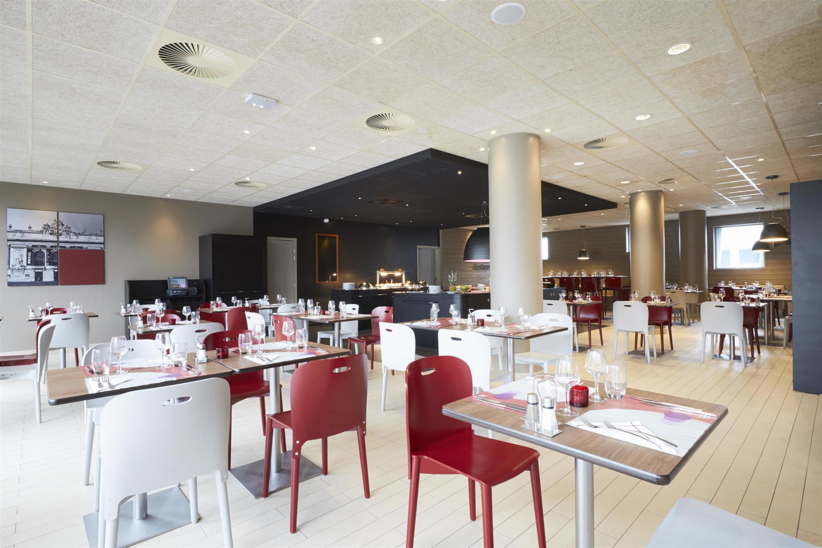 A spacious restaurant with multiple dining areas, including a bar and a separate dining section. Tables are set with red chairs, and the ceiling features recessed lighting and ventilation.