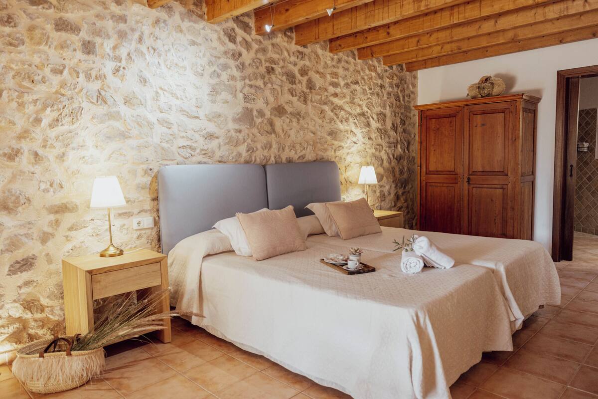 A bedroom with a stone wall, wooden ceiling beams, a bed with two nightstands, a wooden wardrobe, and a woven basket on the floor.
