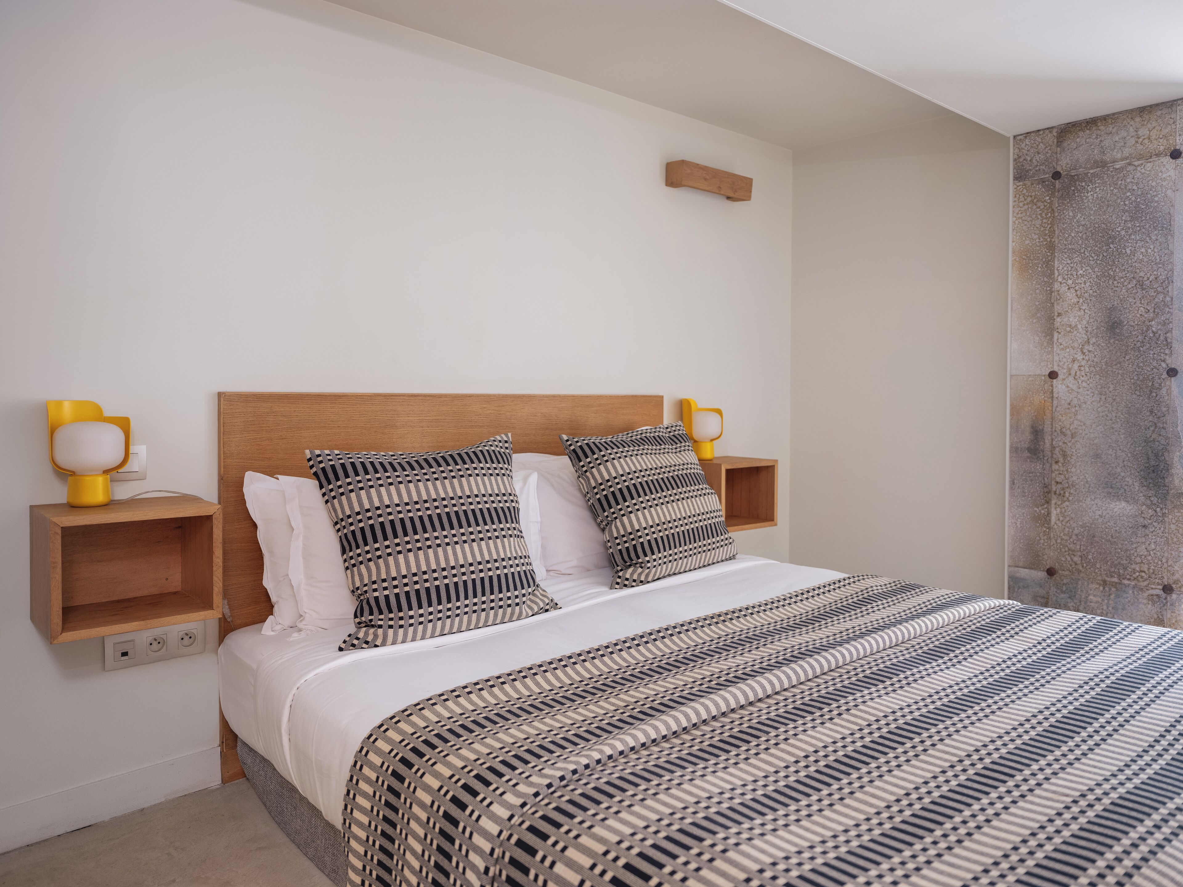 Un dormitorio con cabecera de madera, dos mesitas de noche con lámparas, una colcha estampada y una pared lisa.