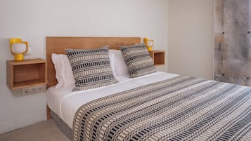 Un dormitorio con cabecera de madera, dos mesitas de noche con lámparas, una colcha estampada y una pared lisa.