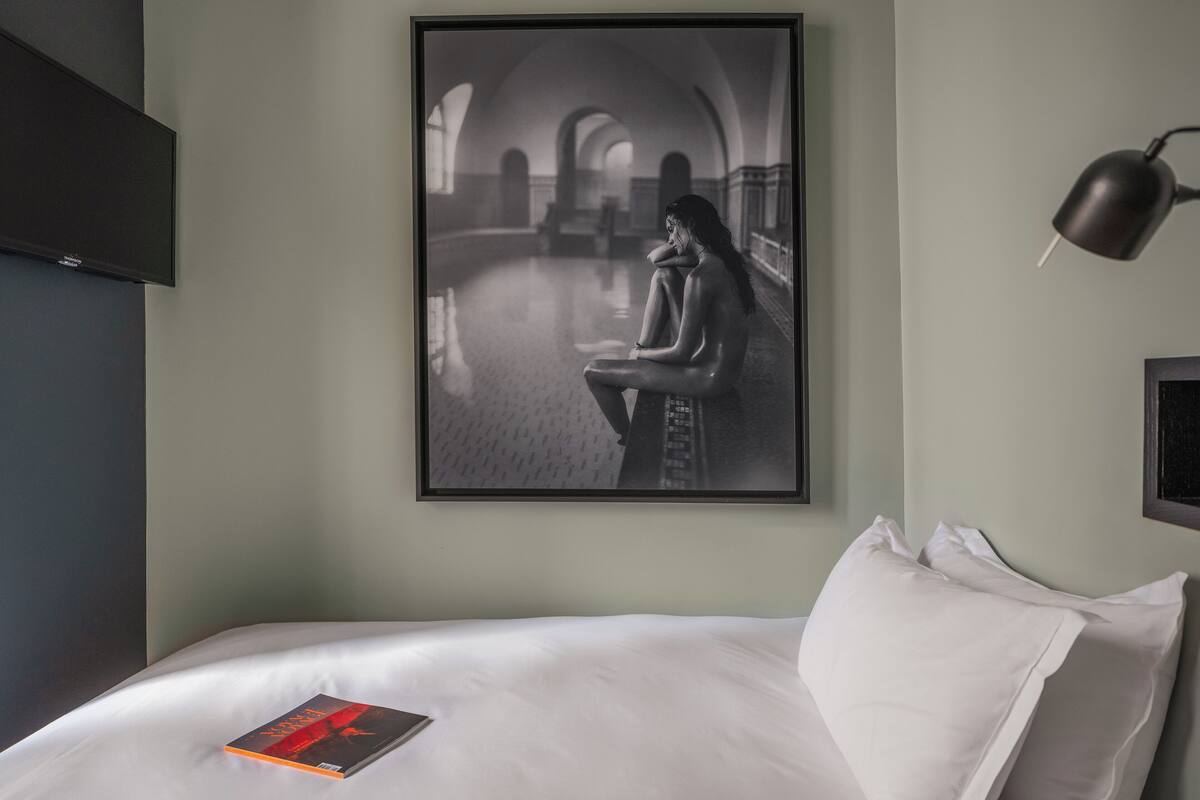 A hotel room with a bed, a framed photograph on the wall, a bedside lamp, and a book on the bed.
