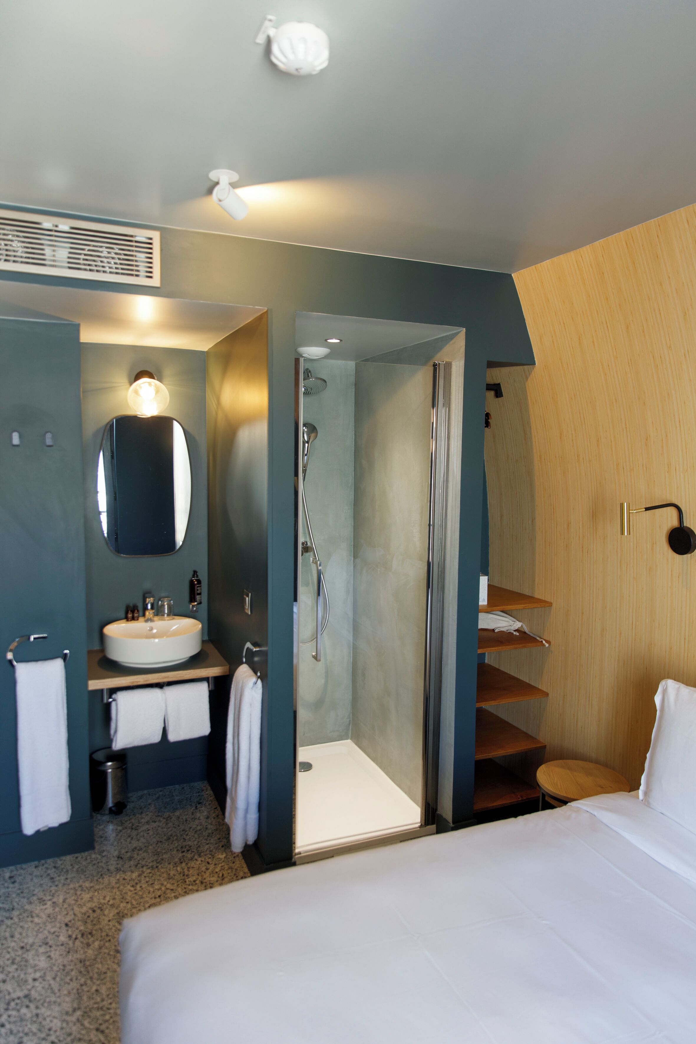 Una habitación de hotel moderna con una cama grande, un baño con lavamanos, espejo y ducha, y un armario de madera.