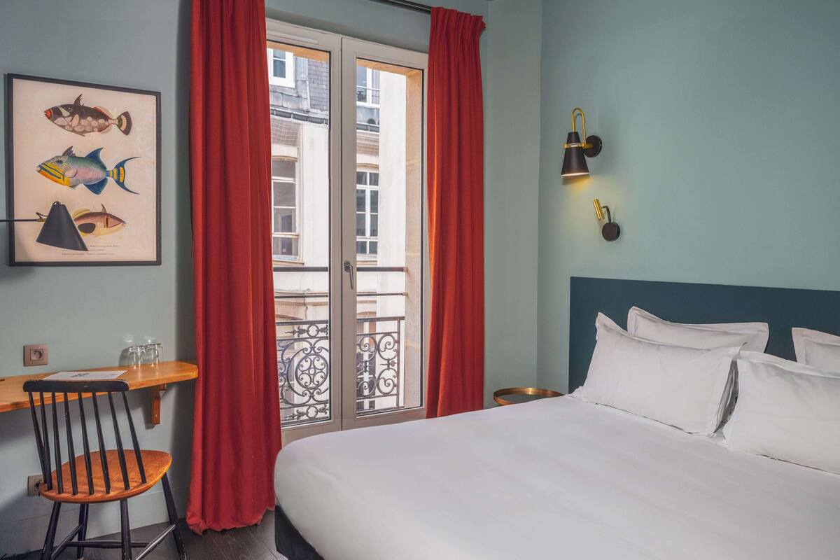 A hotel room with a bed, a desk, a chair, a framed fish illustration, a window with red curtains, and a wall-mounted lamp.