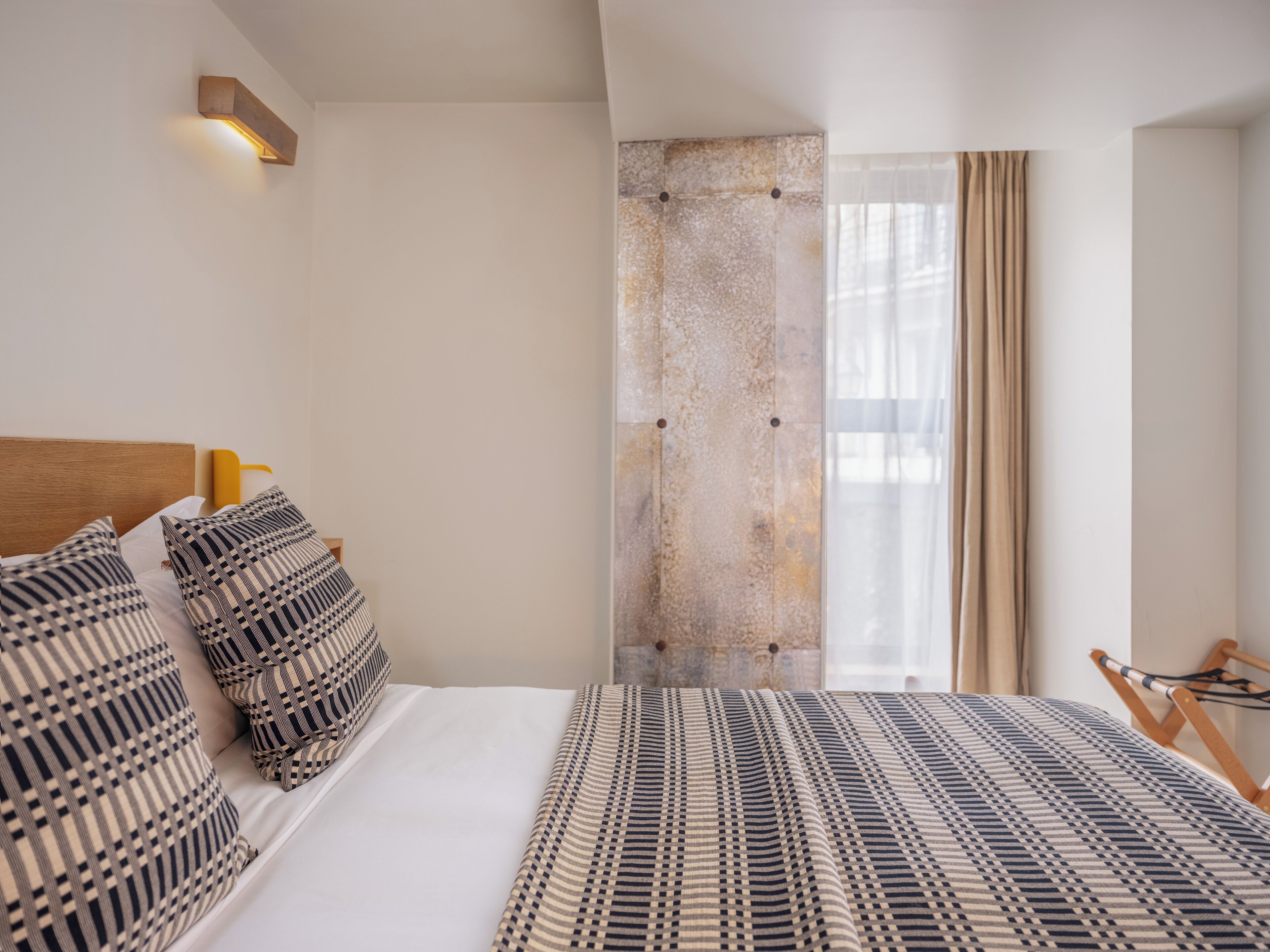 Hotellihuone, jossa on sänky, kuviollisia tyynyjä, sängynpääty, seinään kiinnitetty valaisin, ikkuna verhoilla ja osittainen näkymä puiseen tuoliin.