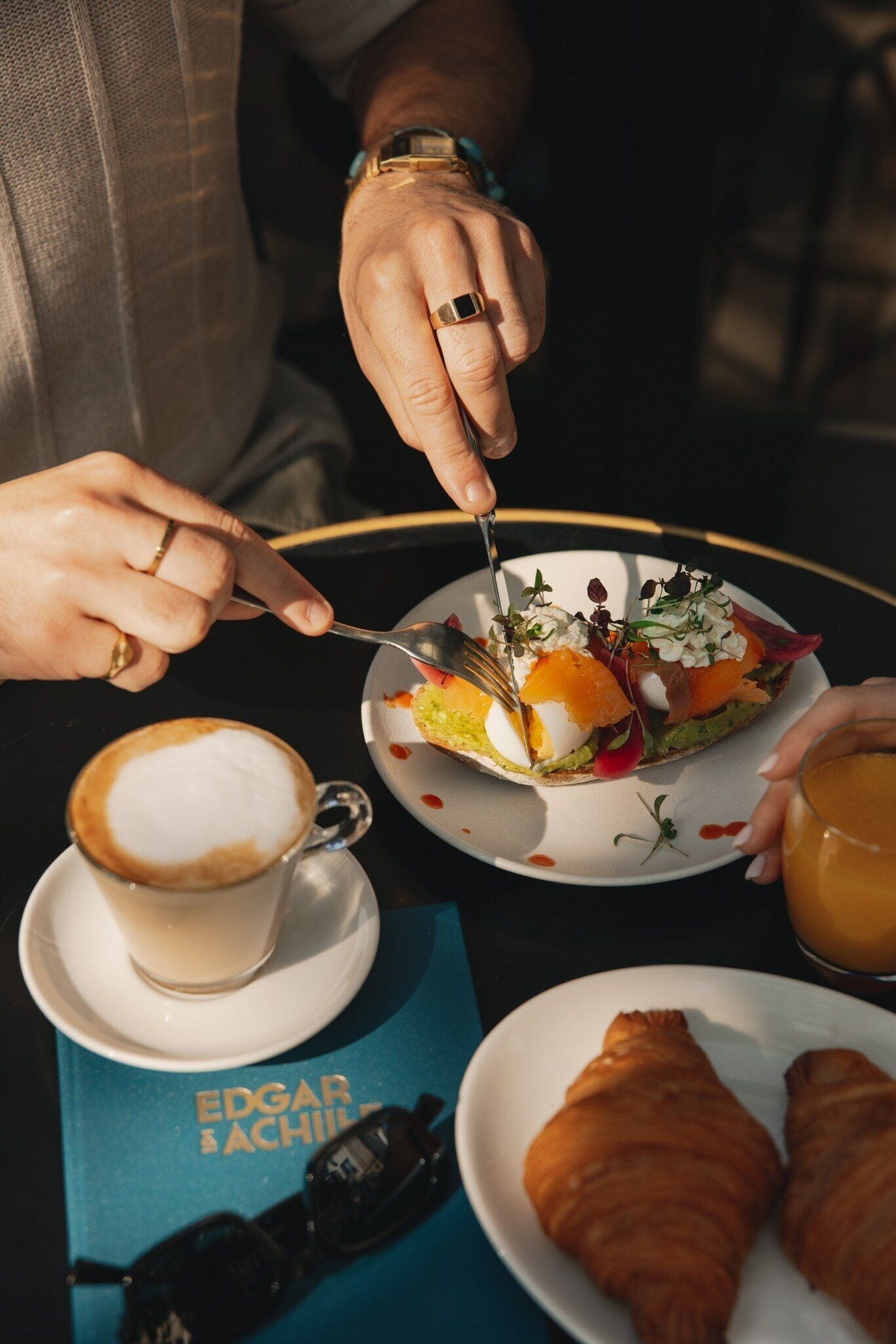 A person dining with a plate of food, a cup of coffee, and a croissant on a table.