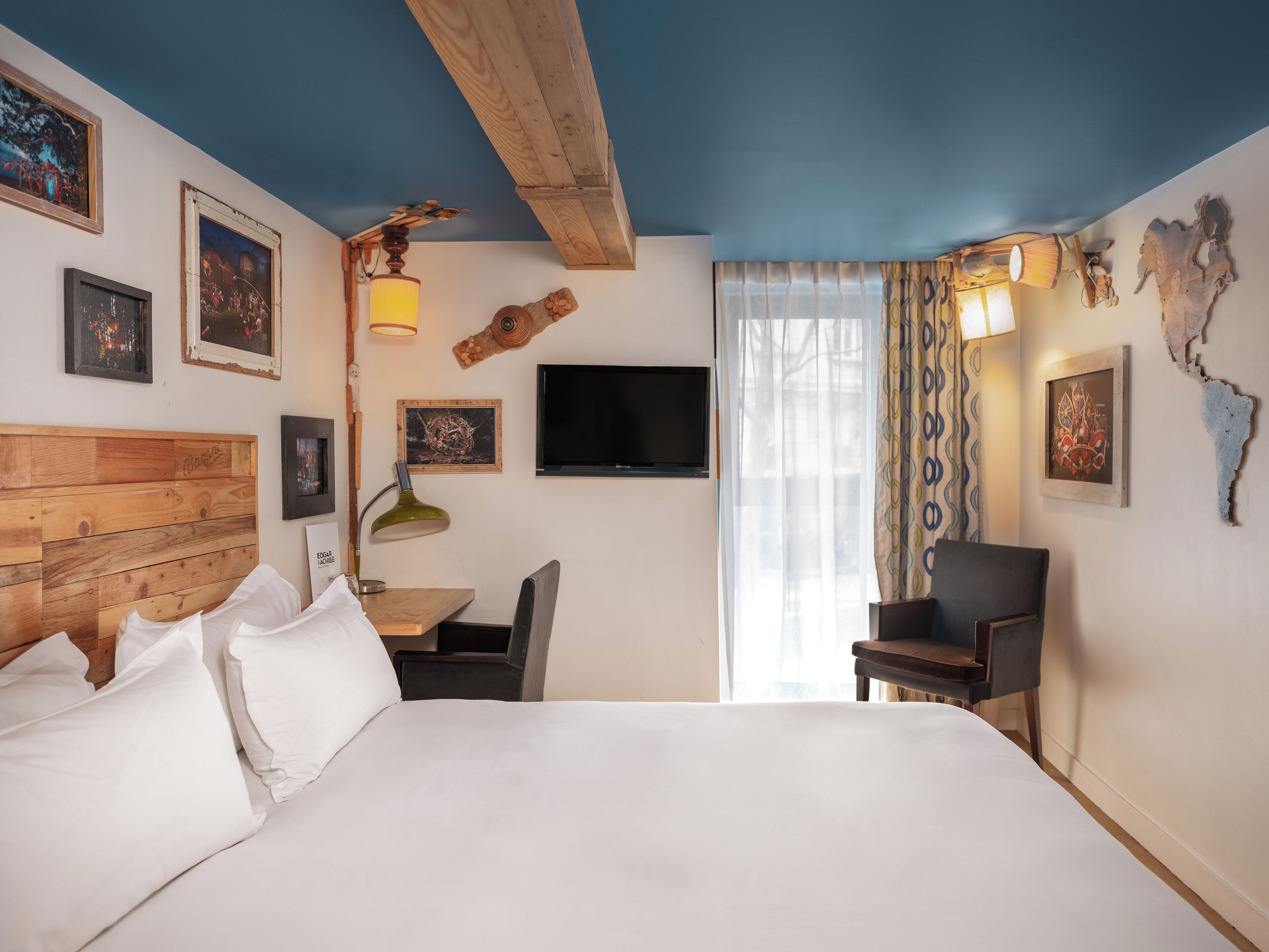 A hotel room with a wooden headboard, a bed with white linens, a desk with a chair, a television, and artwork on the walls.