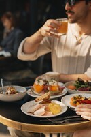 A person holding a glass of beverage at a table with various dishes including a sandwich, salad, and a bowl of food.