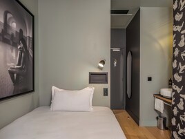 Et moderne hotelværelse med en stor seng, et billede i ramme på væggen, et lille natbord med en lampe, et badeværelse med vask og spejl, samt en bruseniche.