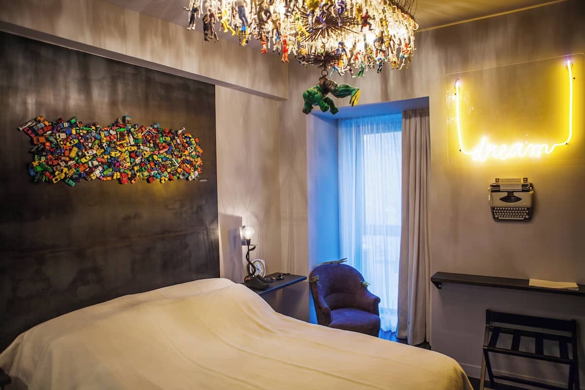 A modern hotel room with a large bed, a desk with a lamp, a chair, a neon sign, and a wall art installation.