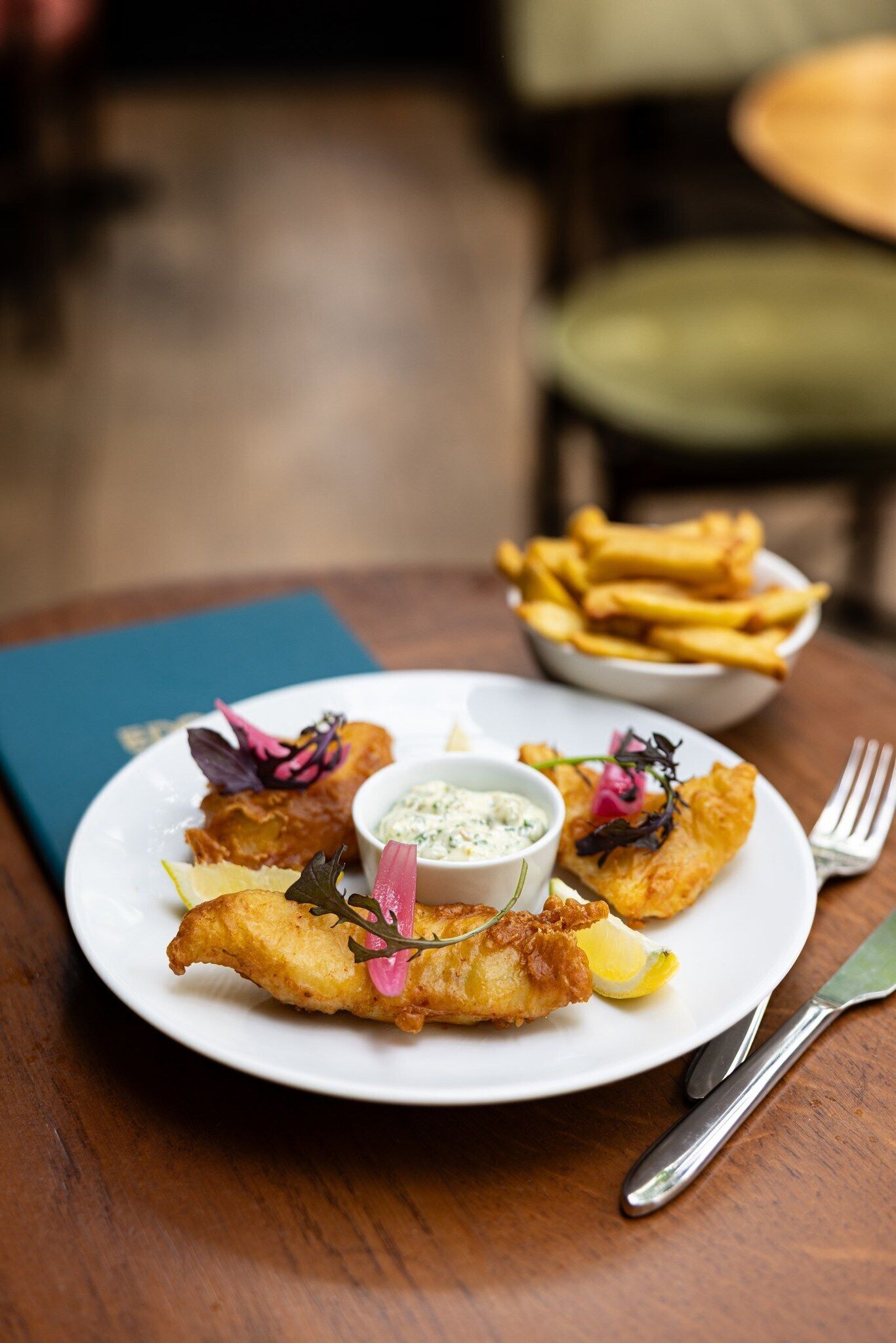 A plate of food with fried items, a bowl of fries, and a small bowl of sauce, on a wooden table with a book and a fork.