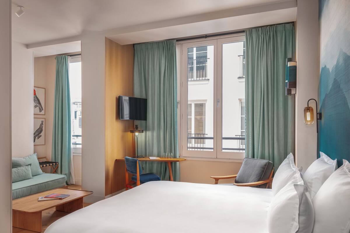 A hotel room with a large bed, a desk with a chair, a television, and a view of the city through the window.