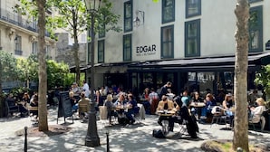 Un área de café al aire libre con mesas y sillas, rodeada de árboles y un edificio con el letrero "EDGAR HOTEL RE/TAURANT".