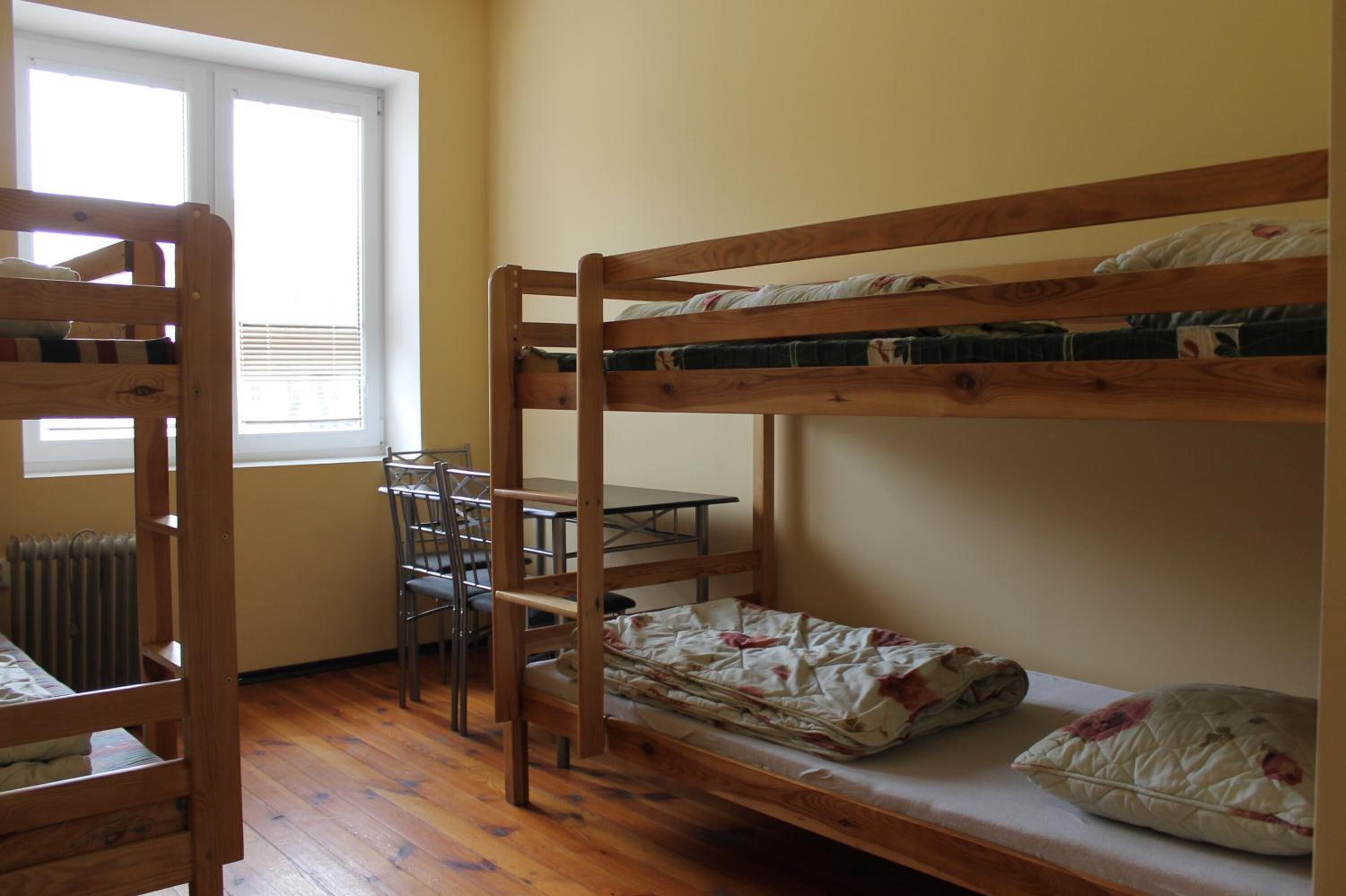 Una camera con un letto a castello, un tavolo, sedie, una finestra e un termosifone.