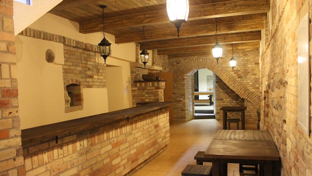 A rustic interior with exposed brick walls, wooden beams, and a stone archway leading to another room. There are wooden tables and benches, and hanging lantern-style lights. - Fortuna Hostel (Vilnius)