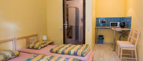 Una habitación pequeña con dos camas, una silla de madera, una kitchenette y la puerta del baño.